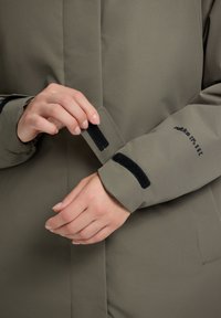 Oliivivärvi takki, millel on sile tekstuur, Velcro mansetiga ja mustad detailid. Lihtne disain, funktsionaalne ja praktiline.