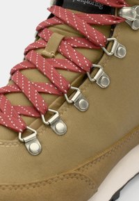 Botte de randonnée marron avec des lacets rouge et blanc, œillets en métal argenté, en toile, et une semelle blanche rembourrée.