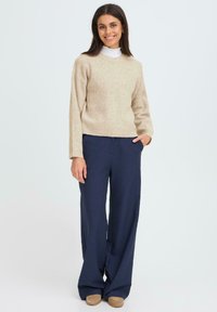 Beżowy prążkowany sweter z długimi rękawami, zestawiony z luźnymi granatowymi spodniami. Model stoi swobodnie z rękami w kieszeniach.