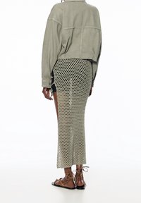 Bijgesneden olijfgroene spijkerjack over een zwart kledingstuk, gecombineerd met een lange beige mesh rok en platte bruine sandalen, gezien van de achterkant.