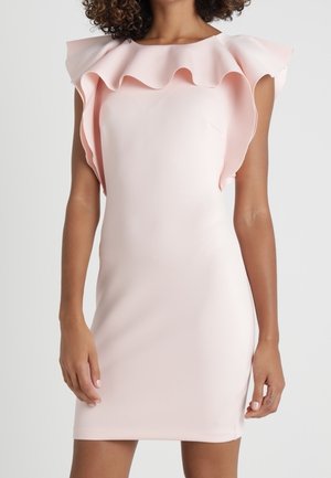 Robe rose pâle moulante sans manches avec grand col à volants et détail aux épaules portée par une personne debout.