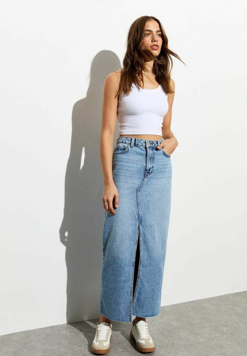 Blauwe denim maxi rok met een voorknip, gecombineerd met een witte tanktop en beige sneakers, tegen een effen witte achtergrond.