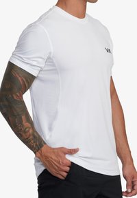 Bílý sportovní triko s krátkým rukávem, prodyšné síťované boční panely a minimální černé logo na hrudi. Hladká textura, přiléhavý design.