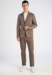 Traje marrón a medida con blazer de un botón y pantalones slim-fit a juego. Llevado sobre una camiseta blanca. Zapatillas blancas completan el look.