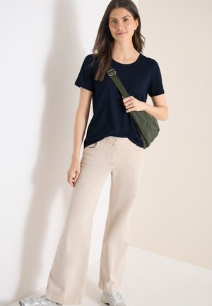 Femme debout contre un mur beige, portant un t-shirt bleu marine, un pantalon ample crème, des baskets blanches et un sac bandoulière vert olive.