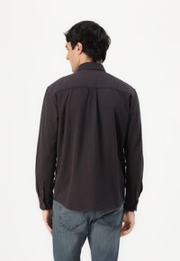 Camicia a maniche lunghe grigio scuro con colletto button-down, dettaglio piega sul retro e polsini con singolo bottone, abbinata a jeans in denim blu chiaro.