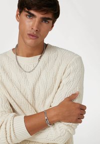 Pull en tricot crème avec un col côtelé, porté en superposition avec une chaîne en argent et un bracelet avec un détail de fermoir. Modèle avec les bras croisés.