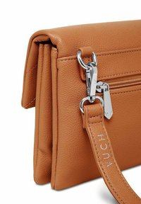 Kabeled žlutě hnědý crossbody batoh s texturovaným povrchem, přední kapsou na zip a stříbrným kováním. Nastavitelný popruh s vyraženým názvem značky.
