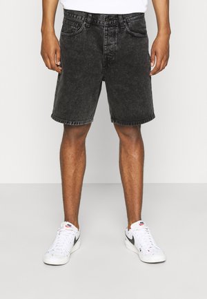 Personne portant un short en jean noir et des baskets blanches Nike, debout devant un fond clair uni.