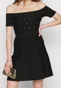 Robe noire décolletée, avec un corps ajusté, des détails texturés et des embellissements dorés, s'évasant en une jupe de style patineuse.