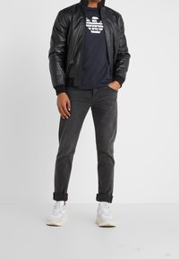 Svart läderbombarjacka, marinblå t-shirt med vit grafik, grå jeans och vita sneakers. Texturerade material, avslappnad passform.