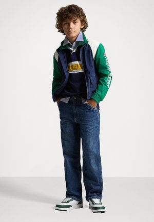 Veste marine et verte sur un polo rayé, associée à un jean bleu et des baskets à accents verts. Comprend un logo sur les manches et un design superposé.