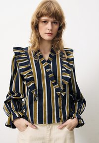 Blouse rayée avec des rayures marine, olive et blanches, présentant des volants le long du col et des manches, une fermeture à boutons et un tissu doux.