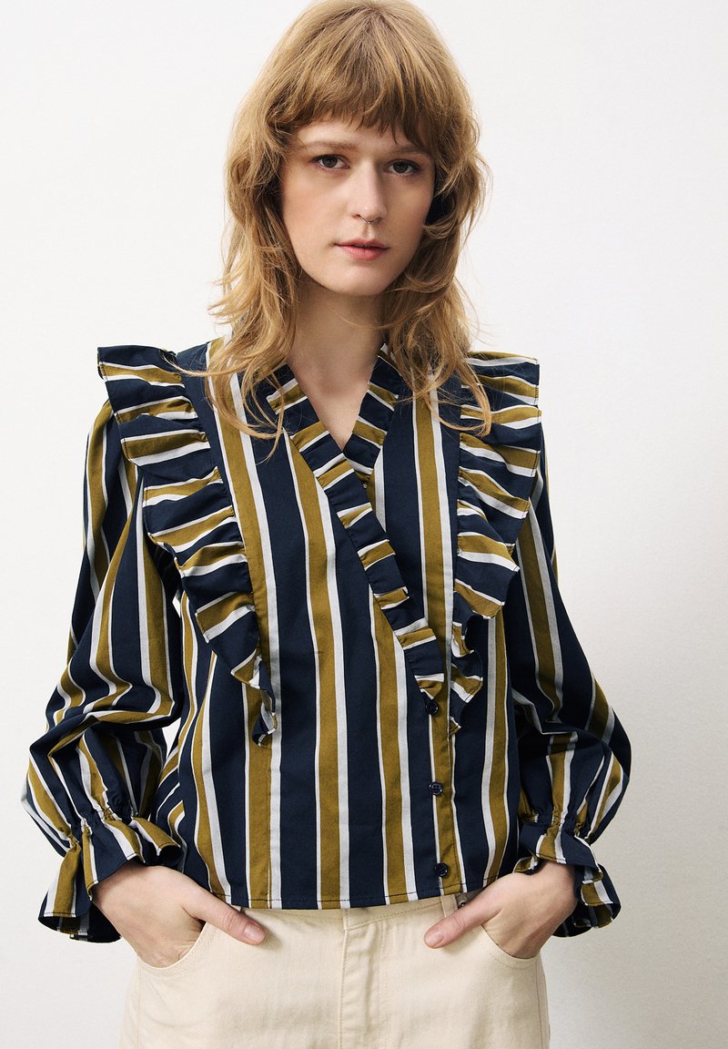 Blouse rayée avec des rayures marine, olive et blanches, présentant des volants le long du col et des manches, une fermeture à boutons et un tissu doux.