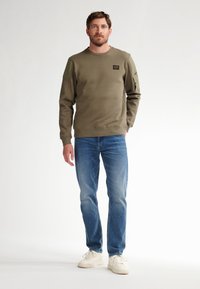 Sudadera verde oliva con acentos texturizados en los hombros, puños acanalados y cuello redondo, combinada con jeans azules y zapatillas de color claro.