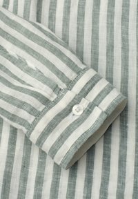 Poignet à manches longues avec bouton blanc sur tissu à rayures verticales gris clair et blanches, probablement un détail de manche de chemise.