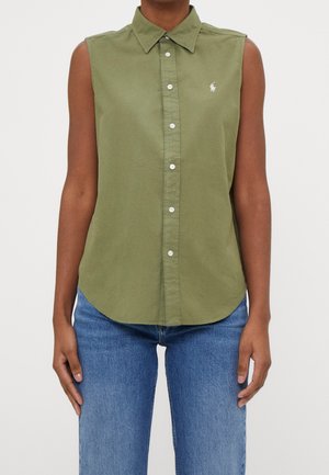 Ärmelloses, olivgrünes Hemd mit Knöpfen und Kragen, getragen von einer Person in blauen Jeans, zeigt Oberkörper und Arme vor einfachem Hintergrund.