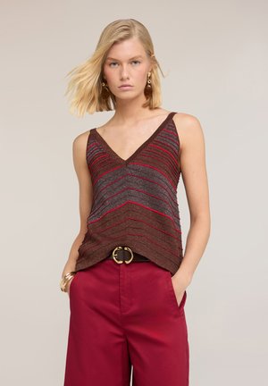 Femme aux cheveux blonds mi-longs, portant un haut sans manches rayé marron et rouge et un pantalon rouge taille haute avec une ceinture noire.