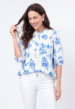 Frau lächelnd, trägt weiße Bluse mit blauem Blumenmuster und Dreiviertelärmeln, kombiniert mit weißen Hosen.