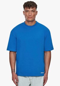 Blå bomulds t-shirt med en afslappet pasform, korte ærmer, rund halsudskæring og et logo detaljeret på ærmet, båret med lyse denim jeans.