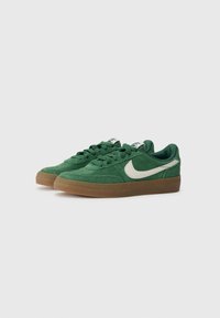 Baskets en daim vert avec un bout arrondi, un Swoosh beige de Nike, des lacets assortis et une semelle en caoutchouc couleur gomme. Un détail de surface texturée est visible.