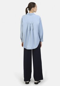 Camicia azzurro chiaro a maniche lunghe con dettaglio a fiocco sulla schiena, vestibilità ampia, orlo arrotondato, abbinata a pantaloni a gamba larga blu navy e scarpe nere.