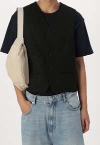 Gilet nero di lana con cinque bottoni, indossato sopra una t-shirt blu navy, abbinato a jeans di denim azzurri e una borsa beige a tracolla.