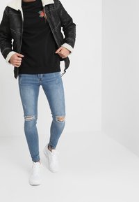 Giacca foderata in finta pelliccia nera, felpa grafica nera, jeans skinny strappati blu chiaro, e sneakers bianche su uno sfondo neutro.