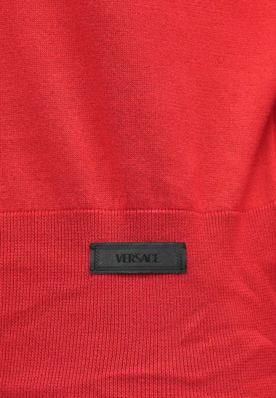 Piros kötött anyag, amelyre egy fekete téglalap alakú címke van varrva, rajta nagybetűkkel a "Versace" márkanév szerepel.