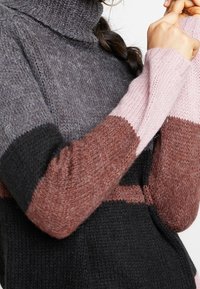 Sweter dzianinowy z kolorowymi blokami w odcieniach szarości, czerni, brązu i różu, noszony przez osobę z warkoczami, trzymającą kołnierz swetra.