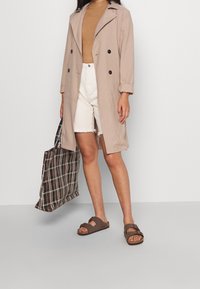 Manteau beige à double boutonnage, haut marron côtelé, short en jean blanc. Porte un sac fourre-tout à carreaux marron, porte des sandales marron.