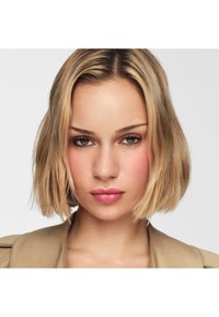 Close-up van een persoon met schouderlang, lichtblond haar en olijfkleurige huid, die natuurlijke make-up draagt met roze lippenstift en een beige jas.