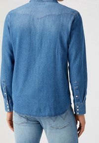 Niebieska koszula dżinsowa z długimi rękawami, zapięciem na guziki i charakterystycznym dla stylu zachodniego karczkiem. Tkanina ma teksturowane wykończenie. Połączona z jasnoniebieskimi dżinsami.