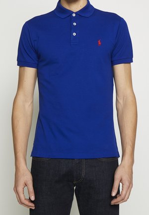 Mann trägt ein eng anliegendes blaues Kurzarm-Poloshirt mit drei Knöpfen und einem kleinen roten Logo auf der linken Brustseite, kombiniert mit dunklen Jeans.