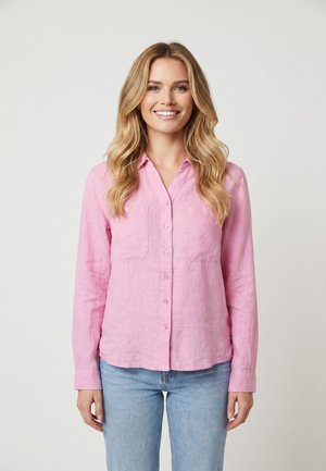 Femme souriante aux longs cheveux blonds portant une chemise rose à boutons et un jean bleu clair, debout devant un fond clair uni.