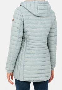 Chaqueta larga acolchada de color gris claro con capucha, cintura ajustada y mangas largas, vista desde la parte posterior en una persona que lleva jeans oscuros.