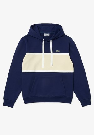 Tummansininen huppari, jossa on beige vaakasuora raita, valkoiset nyörit, etupuolen kengurutasku ja pieni vihreä Lacosten krokotiililogo rinnassa.