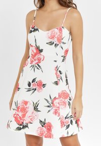 Blommig klänning med vit bakgrund, prydd med stora rosa rosor och gröna blad. Tunna axelband och utställd A-linjeform.