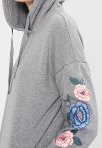 Grå huvtröja med färgglad blommönsterbrodyr på vänsterärm. Tyget verkar mjukt med en avkopplad passform.