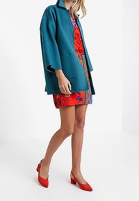 Turkusowy, oversize'owy wełniany płaszcz na czerwonej sukience w wzory, zestawiony z czerwonymi zamszowymi czółenkami. Płaszcz posiada kołnierz i przednie kieszenie. Gładka faktura.