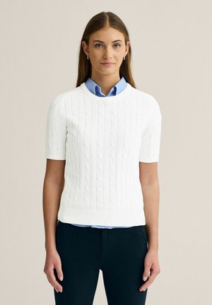 Ung kvinde med glat brunt hår bærer en hvid kortærmet kabelstrikket sweater over en lyseblå skjorte med krave og sorte bukser.