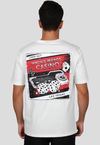 Camiseta de algodón blanca con un estampado gráfico de una escena de casino que incluye dados, cartas de juego y "Wrong Friends' Casino" en rojo y negro.