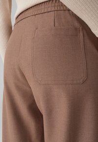 Pantaloni marroni con un tessuto testurizzato, dotati di una tasca posteriore cucita. Vita elastica visibile in cima. Design casual.