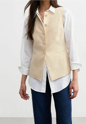 Femme portant un gilet beige boutonné sur une chemise blanche aux manches retroussées et un jean bleu foncé sur un fond blanc uni.