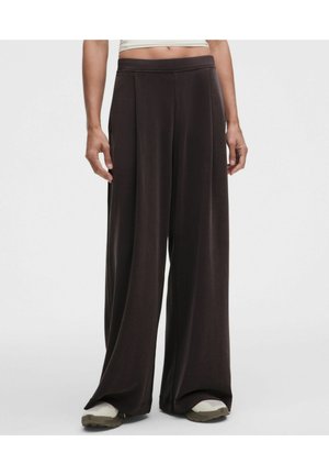Persona que lleva pantalones anchos y holgados de color marrón oscuro combinados con zapatos de color claro, mostrado desde la cintura hasta los pies sobre un fondo liso.