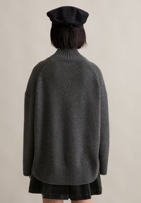 Άτομο που φοράει γκρι ριχτό πουλόβερ με ψηλό λαιμό, μαύρη κοτλέ φούστα και ένα μαύρο υφασμάτινο μπερέ, γυρισμένο μακριά σε έναν απλό φόντο.