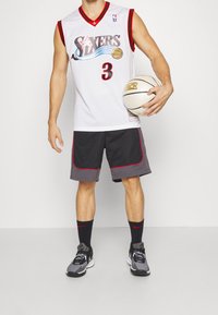 Mies, który nosi białą koszulkę koszykarską Sixers i czarne szorty, trzyma białą piłkę do koszykówki, stojąc na tle jednolitej białej ściany.