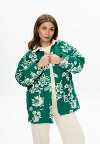 Groene gewatteerde jas met wit bloemenpatroon, voorzien van een kraag, voorzakken en een losse pasvorm. Draagt over een lichte top.