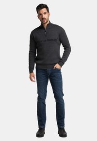 Kohlefarbener Zip-Up-Pullover mit 'bruno banani'-Logo, gerippten Bündchen und Saum, kombiniert mit blauen Jeans und schwarzen Schuhen.