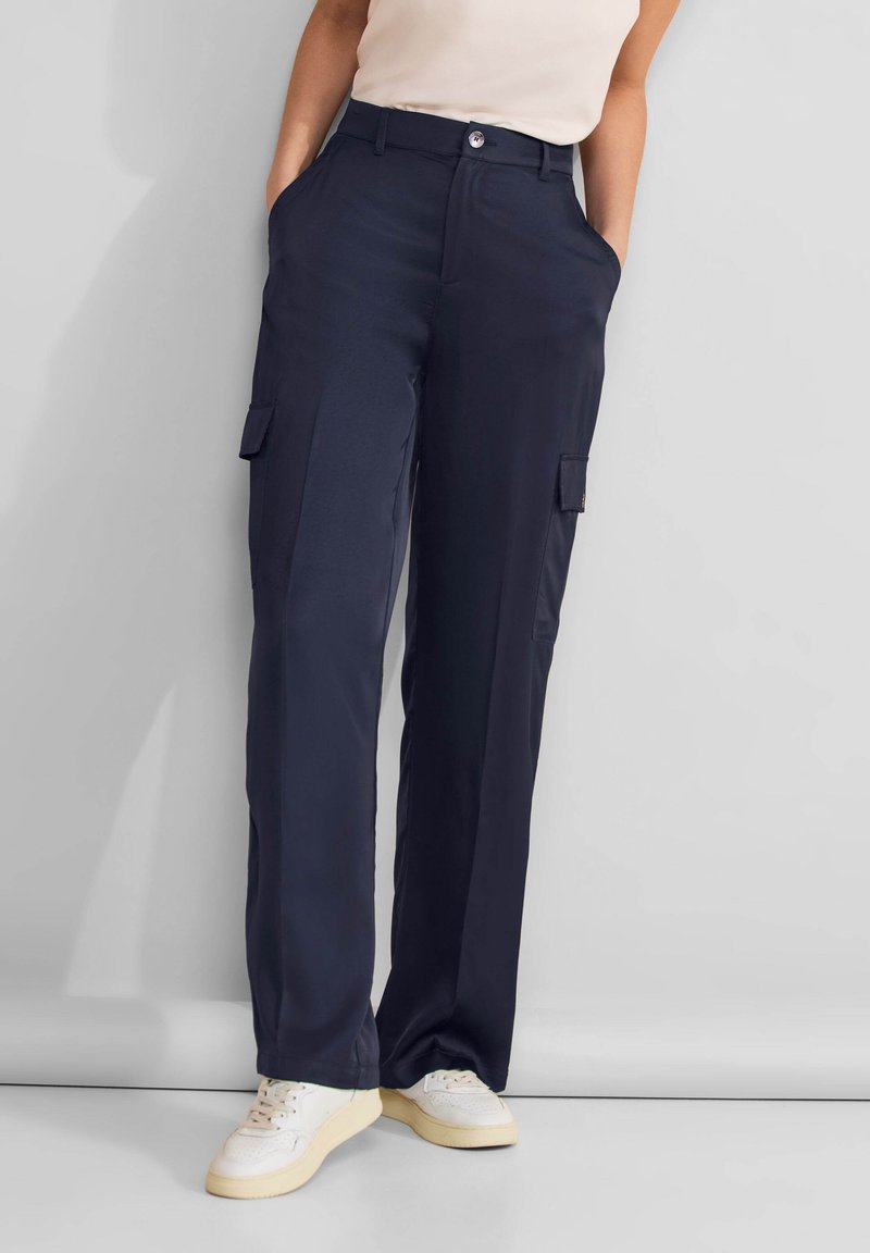 Donna che indossa pantaloni cargo blu navy con tasche laterali e sneakers bianche, in piedi davanti a uno sfondo grigio chiaro uniforme.